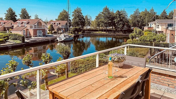 A wooden table with a bottle and glass on a balcony overlooking a canal with boats and buildings.