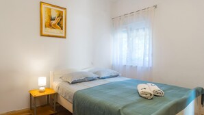 A bedroom with a bed, a bedside table with a lamp, a window with sheer curtains, and a painting on the wall.