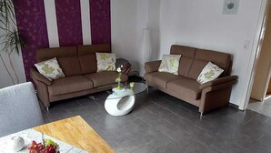 A modern living room with two brown sofas, a glass coffee table, and a wooden dining table with fruit.