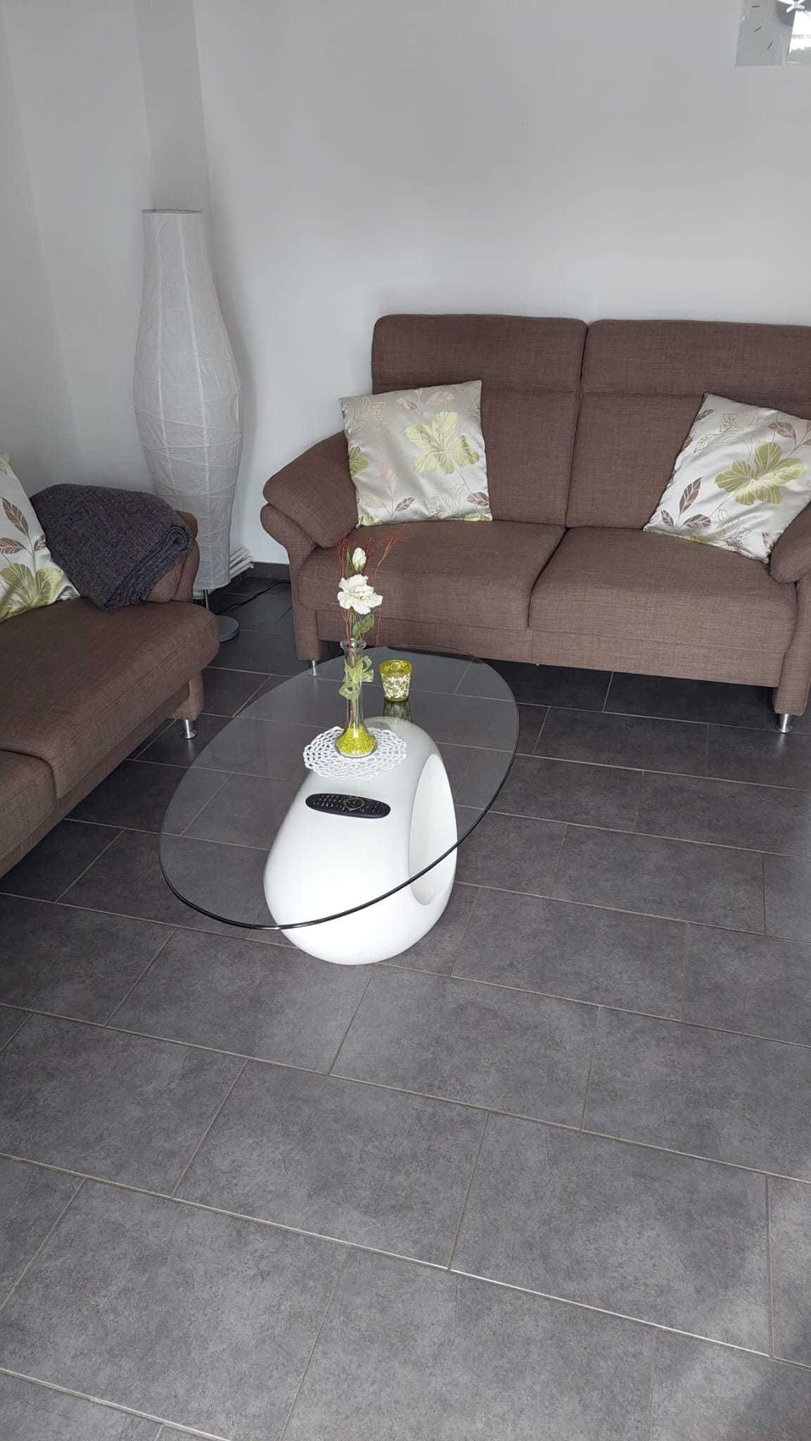 Un salon moderne avec un canapé marron, une table basse blanche ornée d’un vase et de fleurs, et un grand lampadaire blanc.
