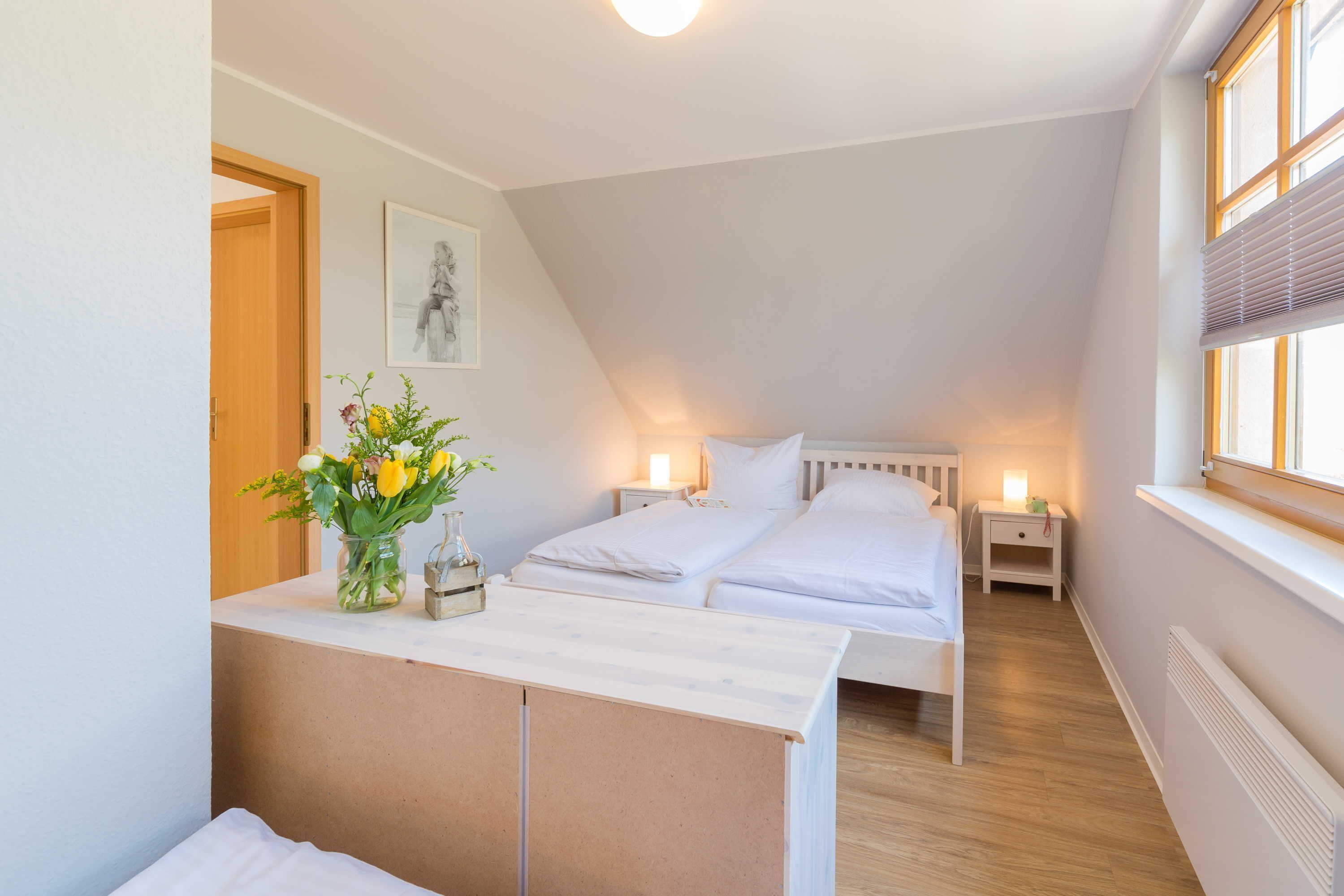 A bedroom with two beds, a bedside table with a lamp, a window with blinds, and a wall-mounted radiator.