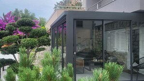 A modern glass-enclosed patio with a stone fireplace, surrounded by greenery and flowering plants. - Beach front apartment in Crikvenica (Crikvenica)