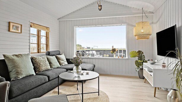 Ein modernes Wohnzimmer mit einem grauen Sofa, einem runden Couchtisch und einem großen Fenster mit Blick ins Freie.