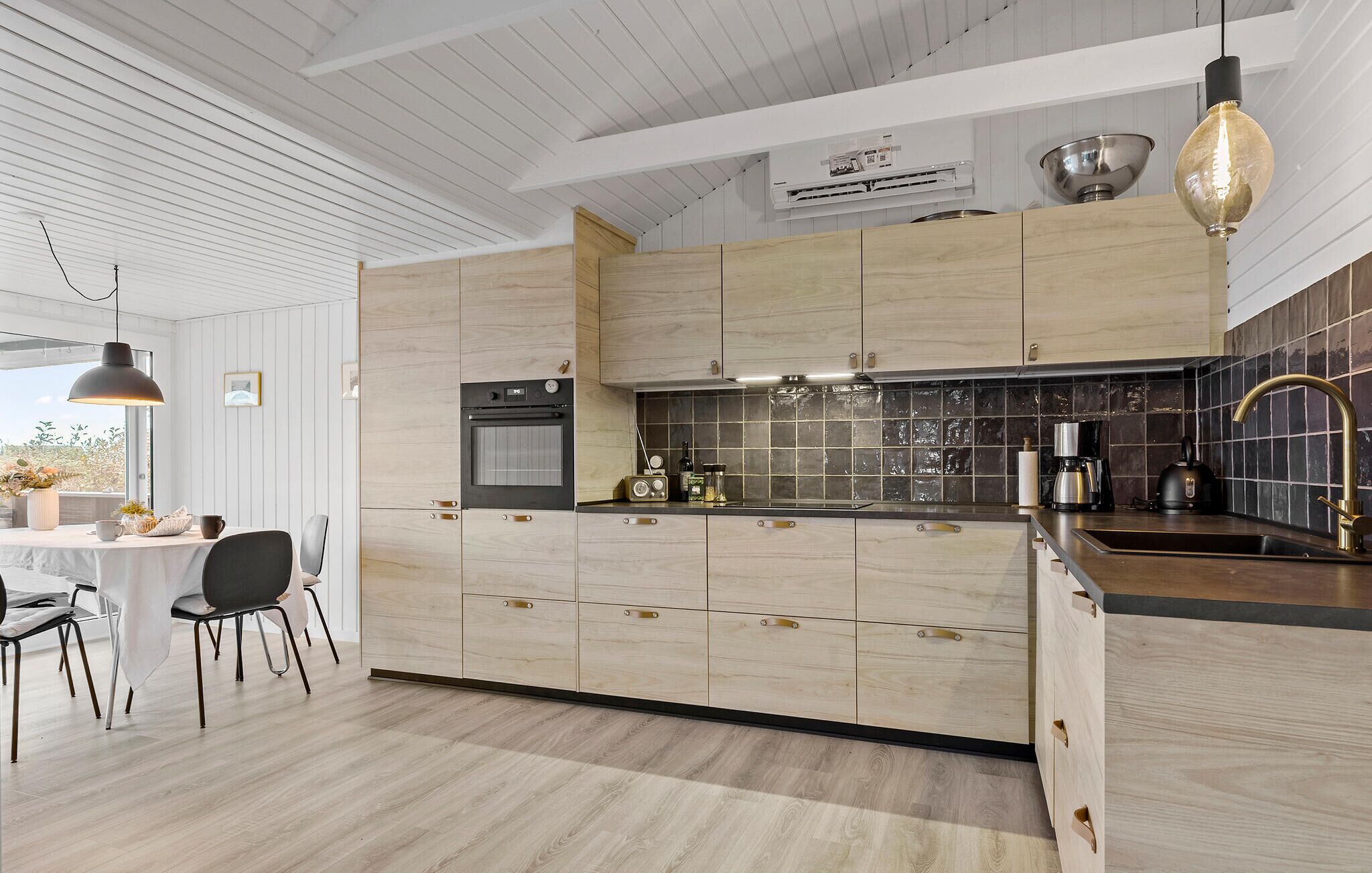 A modern kitchen with light wood cabinets, a built-in oven, a coffee maker, and a sink with a gold faucet.