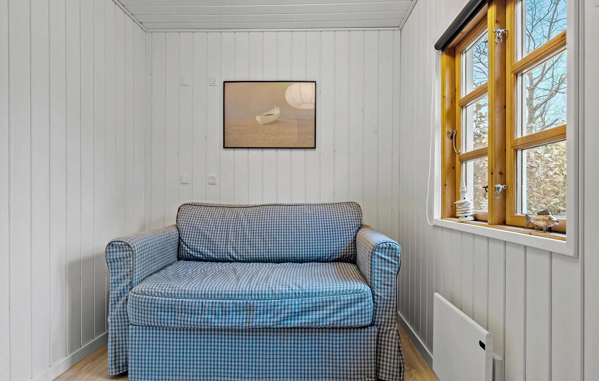 Ein Zimmer mit einem blau karierten Sofa, einem gerahmten Bild an der Wand und einem Fenster mit Blick auf Bäume.