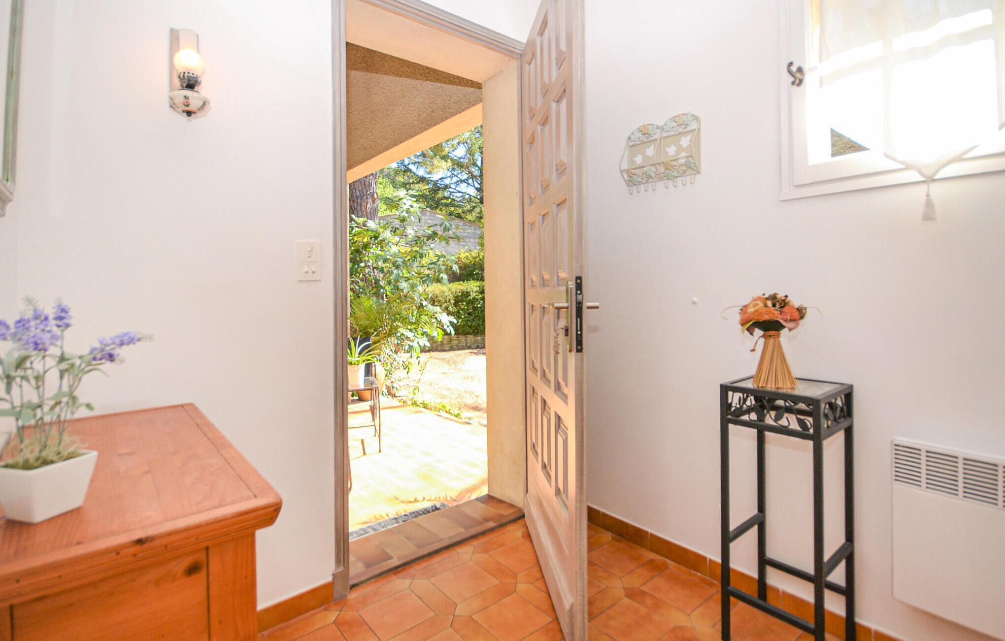 Un couloir avec un banc en bois, une petite table sur laquelle est posé un vase de fleurs, une porte donnant sur un jardin et une fenêtre agrémentée d’un objet décoratif sur le mur.