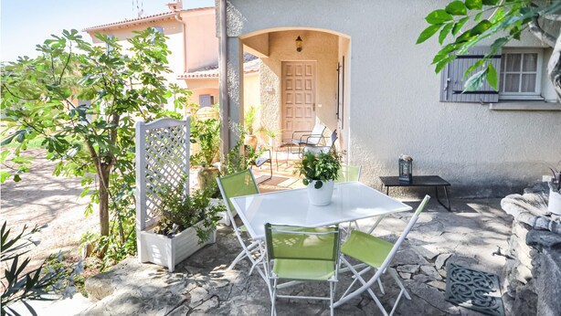 Un patio al aire libre con una mesa blanca y sillas verdes, rodeado de plantas en macetas y un suelo de piedra.