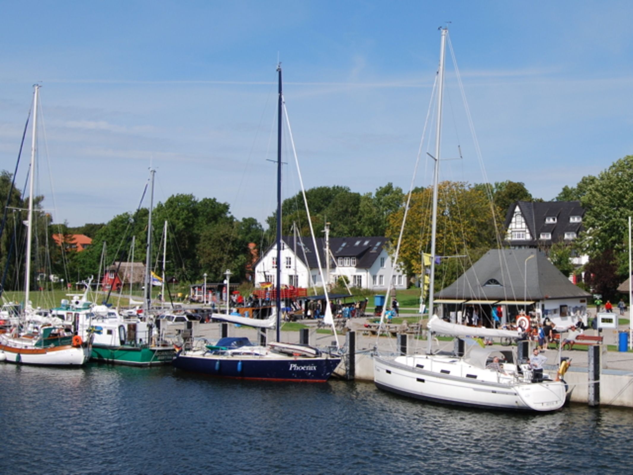 A marina with several boats docked, a white building with a grey roof, and a green area with trees.