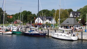 A marina with several boats docked, a white building with a grey roof, and a green area with trees. - Hitthim apartment rental - Apartment harbor view (Insel Hiddensee)