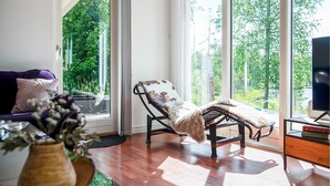 Una habitación con sofá, una silla, una mesita y ventanas amplias con vistas a árboles.