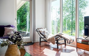 Una habitación con sofá, una silla, una mesita y ventanas amplias con vistas a árboles.