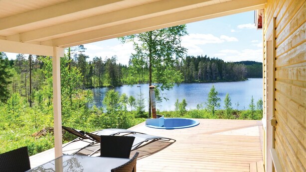 Una terraza techada al aire libre con una mesa y sillas de vidrio, con vistas a un lago y un bosque.