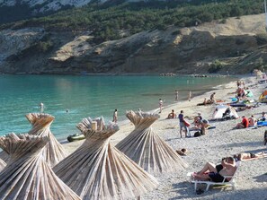 Ein Strand mit Reetdachschirmen, badenden Personen und einer felsigen Hügelkette im Hintergrund.