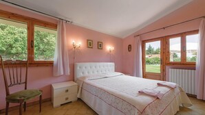 A bedroom with a white bed, a wooden chair, a nightstand, and a window with curtains. - Lovely home in Colle di Compito LU (Colle di Compito LU)