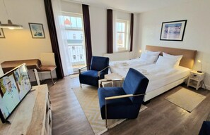Une chambre d’hôtel comprenant un lit, deux fauteuils, une petite table, une table de chevet, un téléviseur et une fenêtre avec des rideaux.