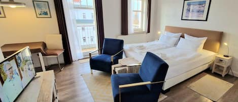Une chambre d’hôtel comprenant un lit, deux fauteuils, une petite table, une table de chevet, un téléviseur et une fenêtre avec des rideaux.