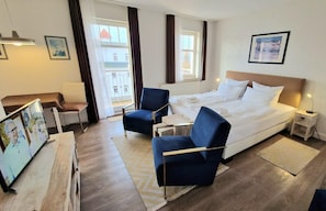Une chambre d’hôtel comprenant un lit, deux fauteuils, une petite table, une table de chevet, un téléviseur et une fenêtre avec des rideaux.