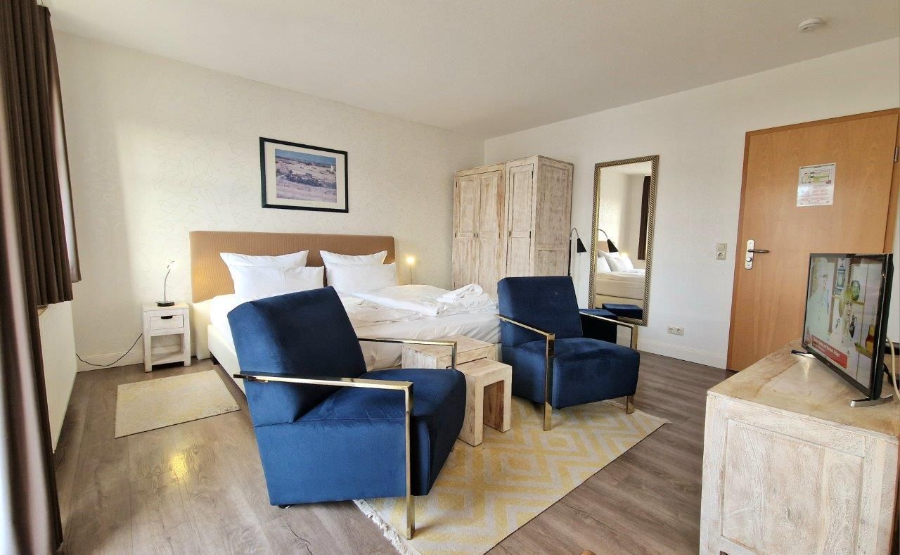Hotellihuone, jossa on sänky, kaksi sinistä nojatuolia, puinen sivupöytä, pieni valkoinen yöpöytä, peili, vaatekaappi ja televisio, joka on sijoitettu kaappiin.