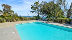 A clear blue swimming pool surrounded by a paved deck, with a view of the ocean and various trees and shrubs. - Gorgeous apartment in Moriani Plage (Moriani Plage)