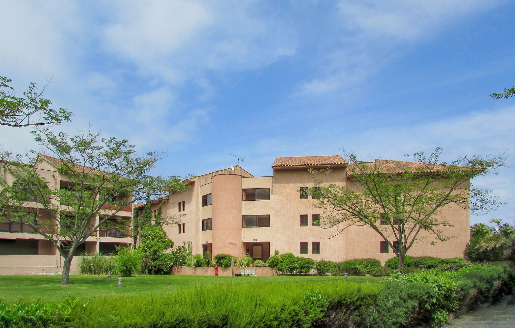 A multi-story residential building with a flat roof, surrounded by well-maintained greenery and a clear sky.