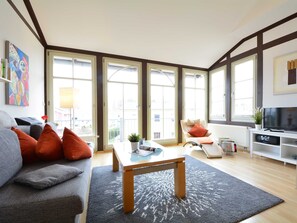 Una sala de estar moderna con un sofá, una mesa de centro y un televisor. Amplios ventanales ofrecen luz natural y vistas al exterior.