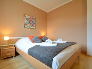 Una cama bien hecha con una manta gris, dos almohadas y toallas enrolladas sobre un piso de madera.