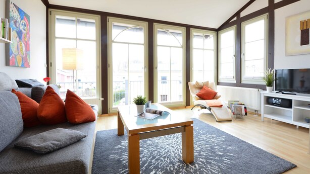 Una sala de estar moderna con un sofá, una mesa de centro y un televisor. Amplios ventanales ofrecen luz natural y vistas al exterior.