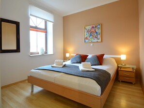 A modern bedroom with a large bed, two bedside tables, a window with blinds, and a colorful abstract painting on the wall.