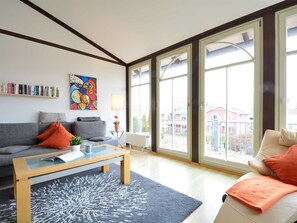 A modern living room with a grey sofa, a wooden coffee table, and a blue rug. There is a bookshelf with books and a colorful painting on the wall.