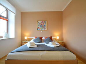 A neatly made bed with a gray blanket and two white towels, a colorful abstract painting on the wall, and two bedside lamps.