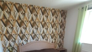 Een slaapkamer met een behang, een bed, een nachtkastje, een bankje en een raam met gordijnen.