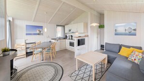 A spacious living area with a grey sofa, a dining table with chairs, a kitchen with white cabinets, and a large window. - Friedrichskoog-Deichblick 20 (Friedrichskoog)
