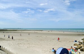 Une vaste plage où l’on voit des gens se promener et jouer, une tente installée, et une mer calme en arrière-plan.