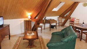 Une pièce mansardée chaleureuse avec un plafond en bois, un canapé vert, une petite table recouverte d’une nappe en dentelle, une lampe, un téléviseur et une table à manger avec des chaises.