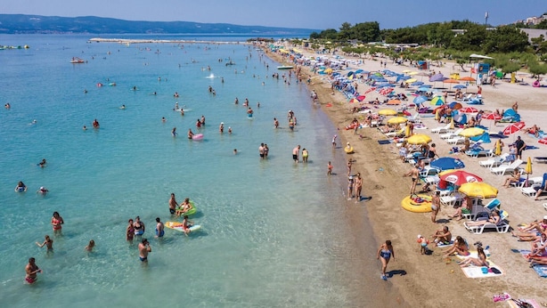Een druk strand met helder water, veel parasols en mensen die van alles doen.