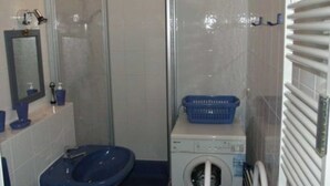 A bathroom with a glass shower enclosure, a sink, a washing machine, and a blue basket on the machine. - FeWo Möwennest / Felstehausen GM 69252 - FeWo Möwennest (Graal-Müritz)