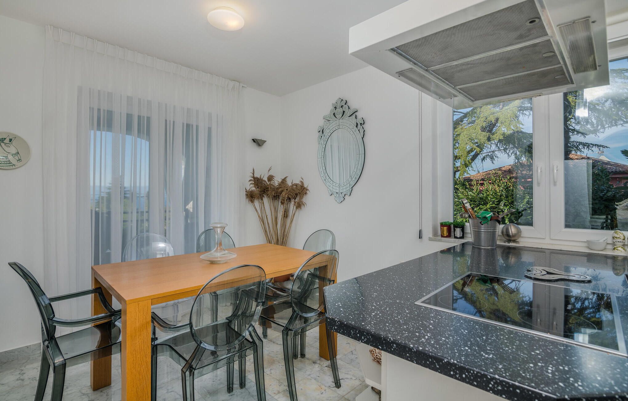 A modern kitchen with a wooden dining table, clear glass chairs, a black speckled countertop, and a large window with a view of greenery.