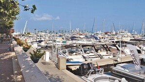 Uma marina com vários barcos atracados, um restaurante chamado "La Trattoria" à esquerda e um céu azul claro ao fundo.