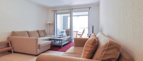 A living room with a sofa, a coffee table, and a chair. There is a window with curtains, a lamp, and a view of the outside.
