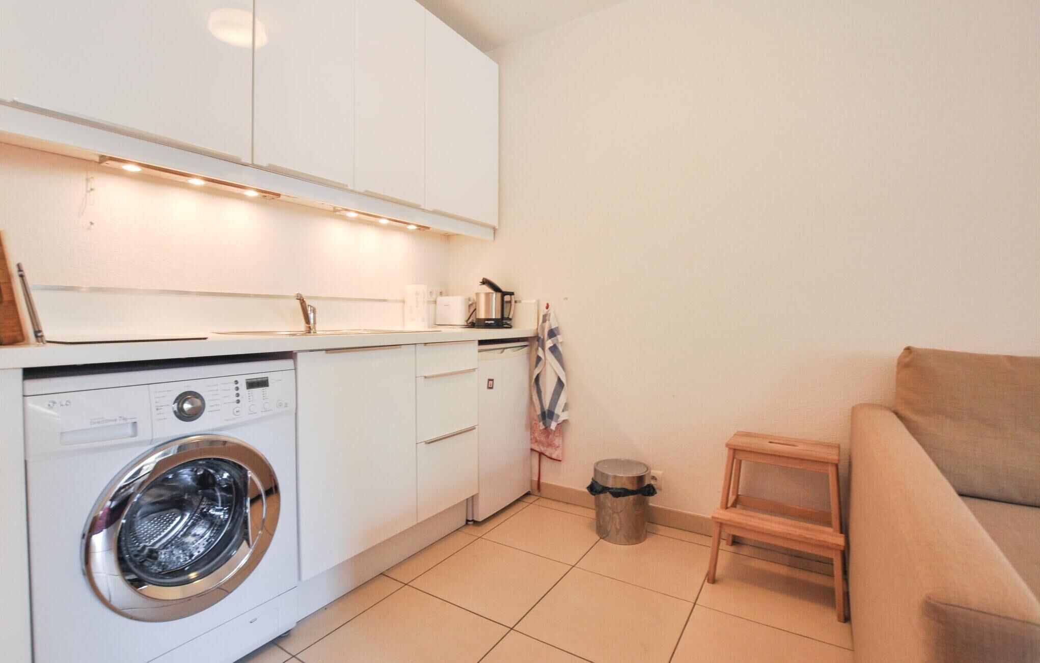 Cocina compacta con lavadora, un taburete de madera pequeño y un cubo de basura.
