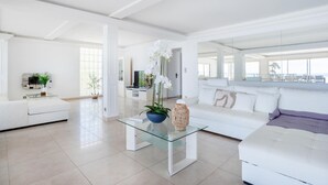 Un salon spacieux avec un canapé blanc, une table basse en verre et une grande fenêtre donnant sur la mer.