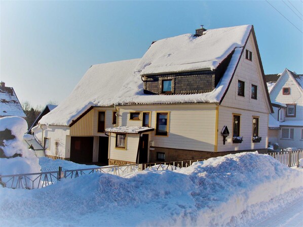 A two-story house with a sloped roof covered in snow, a visible chimney, and a small porch area.