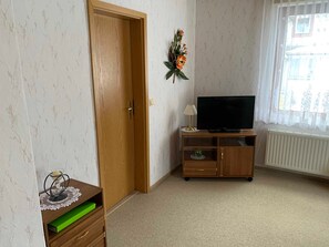 A room with a wooden door, a television on a wooden stand, a radiator, and a window with lace curtains. - FW 2 (in the house) - Apartments Rießling (Elend)