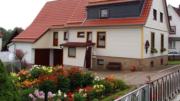 Una casa a due piani con tetto in tegole rosse, un portico e un giardino con diversi fiori.