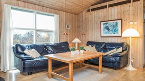Una habitación acogedora con paredes y techo de madera, un ventanal con cortinas, un sofá de cuero negro, una mesita de centro de madera y una lámpara de pie.
