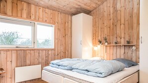 Un dormitorio con techo de madera, un ventanal grande, cama con ropa de cama azul, mesita de noche y estante con plantas.