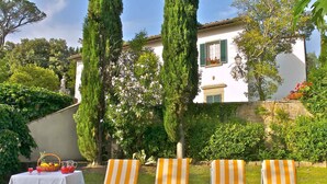 Eine Terrasse mit drei gestreiften Liegestühlen, einem Tisch mit einer Fruchtkorb und einer Kanne, und ein weißes Gebäude mit grünen Fensterläden im Hintergrund.