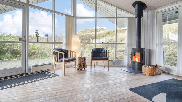 Une pièce spacieuse avec de grandes fenêtres, un poêle à bois noir, deux fauteuils en cuir et un panier en osier.