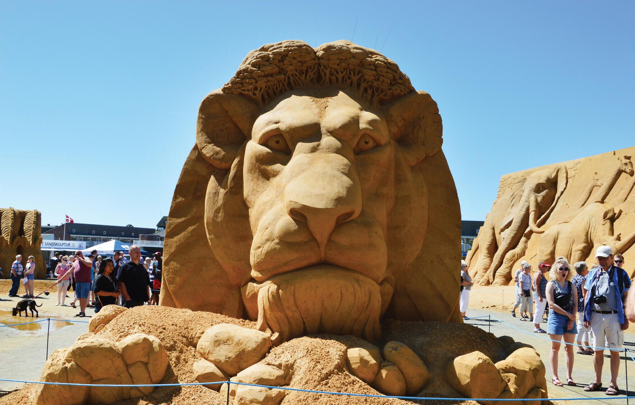 Una gran escultura de sorra amb el rostre d'un lleó, envoltada de petites escultures, com ara una girafa, en un esdeveniment a l'aire lliure sobre la sorra.