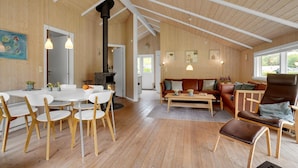 A spacious room with a high ceiling, wooden walls, a dining area with white chairs, a sofa with cushions, and a fireplace. - Awesome home in Juelsminde with sauna (Juelsminde)